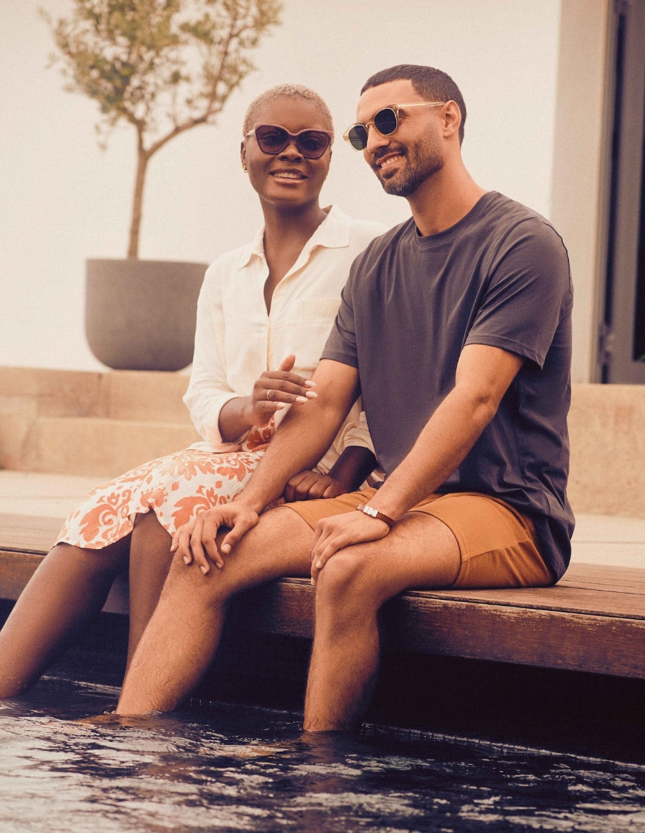 Paar sitzt mit Sonnenbrillen am Pool und lässt die Beine ins Wasser baumeln.